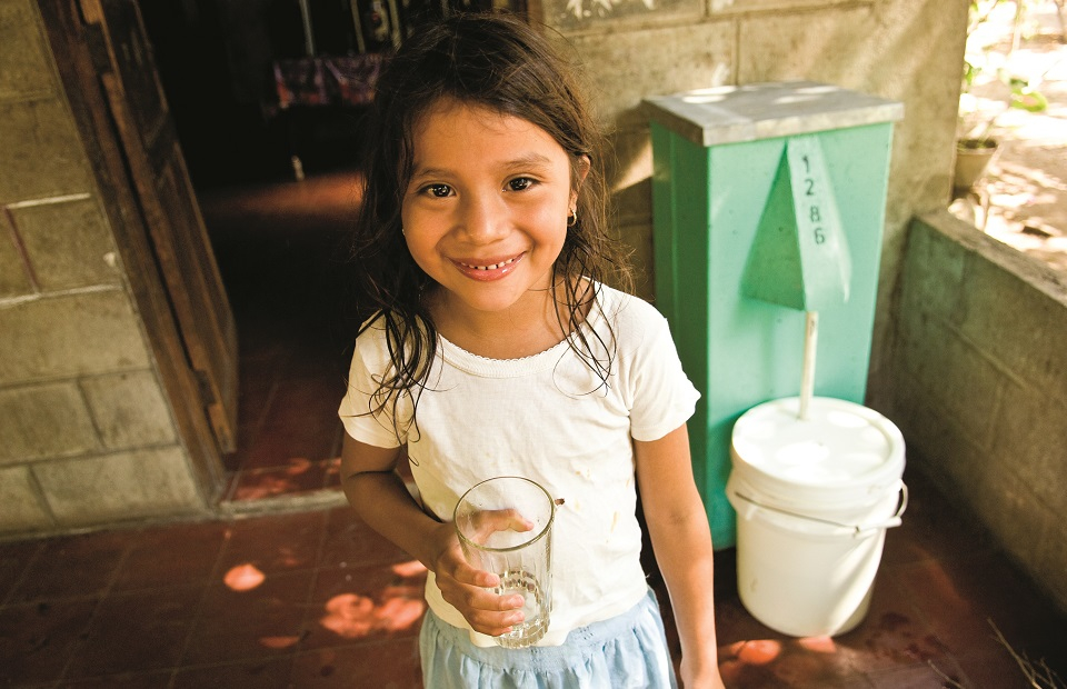 Ein Mädchen hält ein Glas sauberes Wasser in der Hand, hinter ihr der Bio-Sand-Wasserfilter